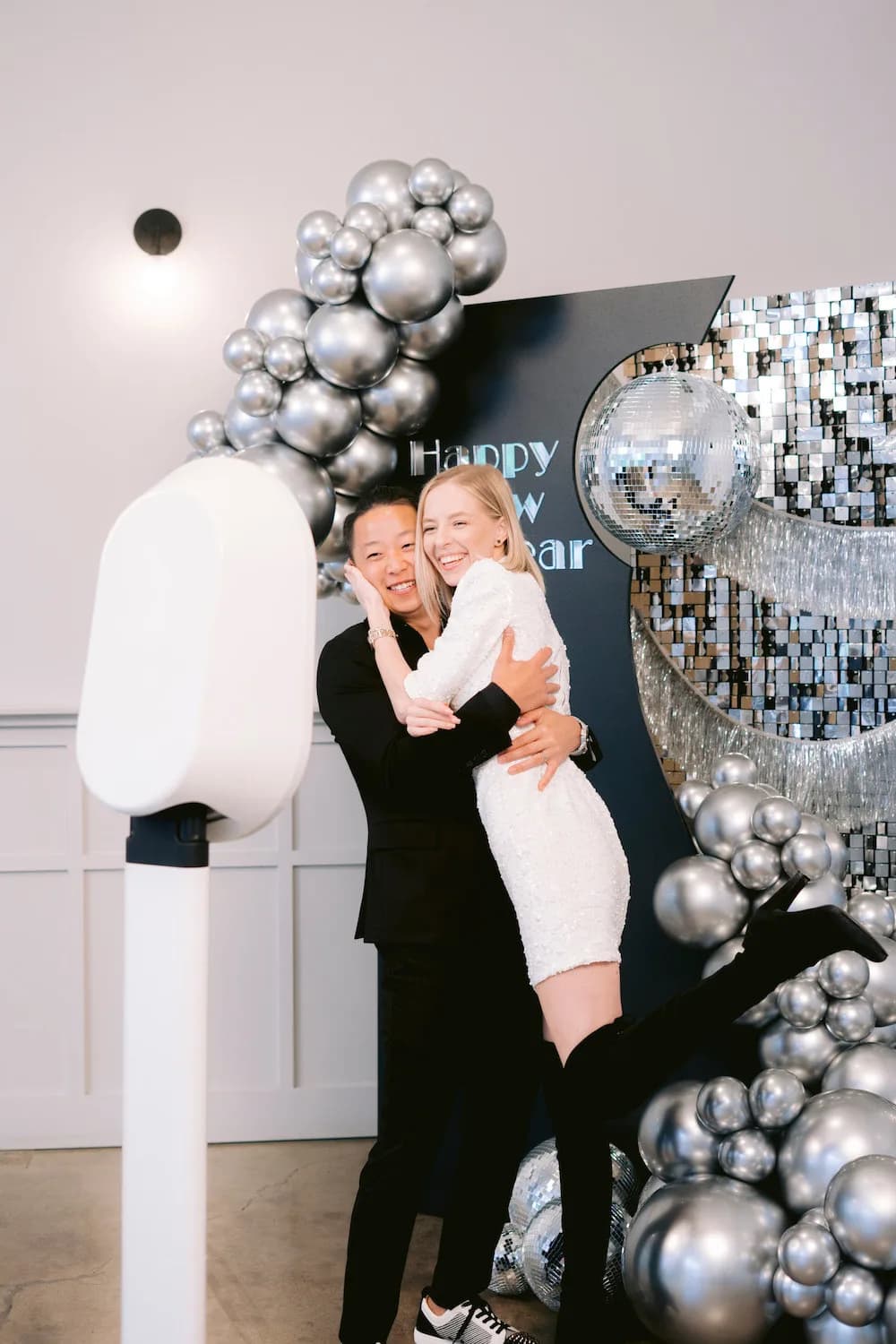 A couple hugging beside a white open-air photo booth, with a silver sequin and balloon backdrop behind them