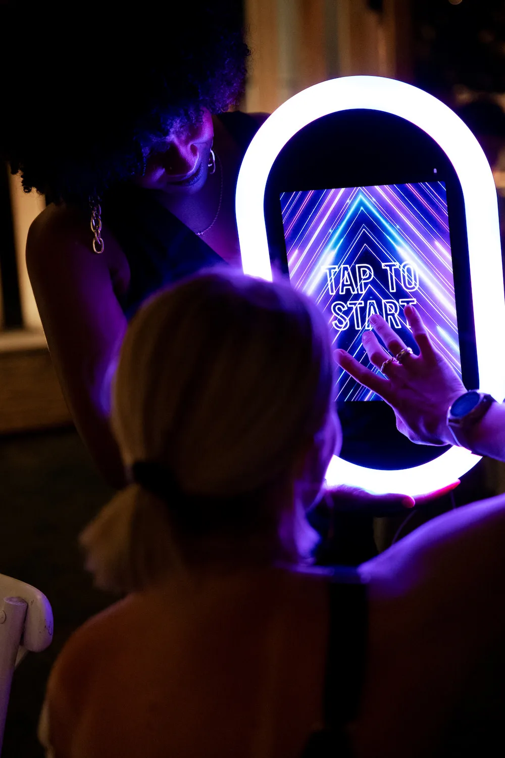 Guests at an open-air photo booth, one person tapping the ring-lit screen that says tap to start