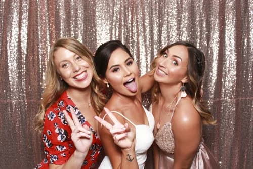 Three guests posing in front of a rose gold sequin photo booth backdrop